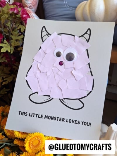 A kids craft features a torn paper monster outlined on paper, covered with pink torn paper pieces, two googly eyes, and a gem nose. Text below reads THIS LITTLE MONSTER LOVES YOU! Flowers are beside the artwork.