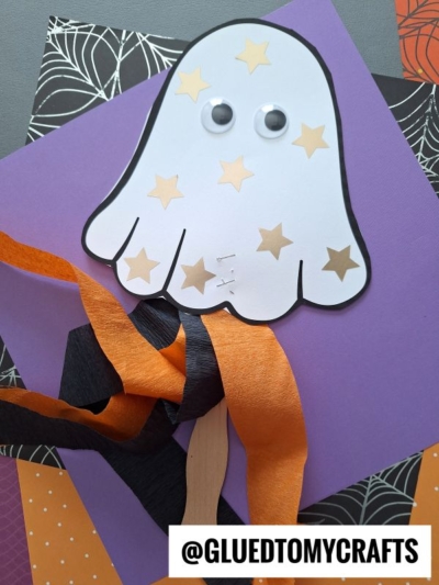 A paper ghost wand craft with googly eyes, gold star stickers, and orange and black streamers on a popsicle stick, displayed on layered Halloween-themed papers. @GLUEDTOMYCRAFTS is written at the bottom.