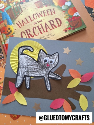 A children’s Halloween craft featuring a cat on tree branch made from black-and-white paper with googly eyes, surrounded by colorful paper leaves, with the book Halloween in the Orchard in the background.