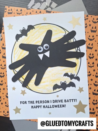 A Halloween card featuring a handprint batty design, with black handprint wings, googly eyes, and fangs perched on a yellow moon—surrounded by stars and bats. The playful text reads, FOR THE PERSON I DRIVE BATTY! HAPPY HALLOWEEN! @GLUEDTOMYCRAFTS.