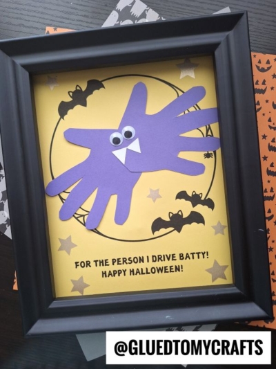 A framed Halloween craft featuring two purple handprint batty wings with googly eyes and fangs on yellow paper, decorated with bats and stars. Caption reads, “For the person I drive batty! Happy Halloween!”.