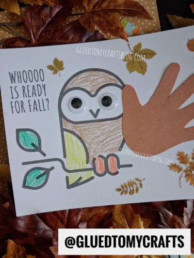 A fall-themed craft features a charming handprint owl drawn on paper, with googly eyes and a handprint for a wing. Text reads “Whoooo is ready for fall?” Leaves and craft supplies are scattered around.