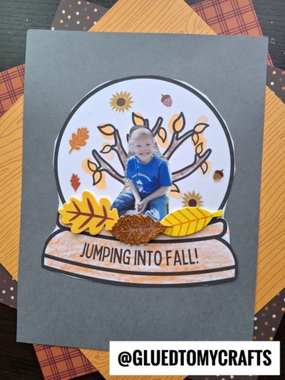 A handmade Fall Snow Globe Keepsake craft features a child’s photo on a paper snow globe adorned with autumn leaves and the text JUMPING INTO FALL! on a dark background. The @GluedToMyCrafts watermark is at the bottom.