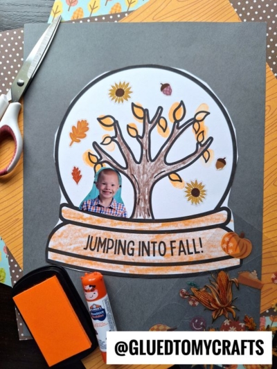 A Fall Snow Globe Keepsake craft features a drawing with a tree, autumn leaves, and a cut-out photo of a smiling child. Scissors, glue, and an ink pad surround the art. Text on the globe reads, “JUMPING INTO FALL!”.