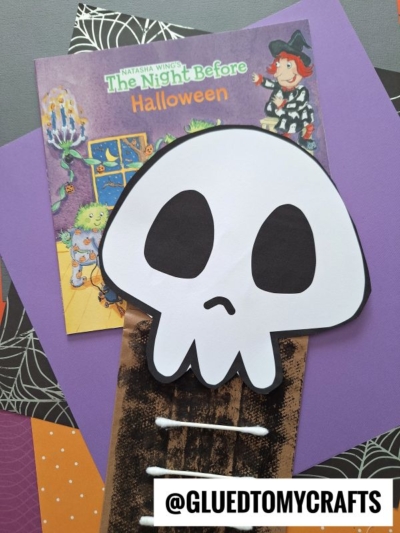 A Halloween book, purple paper, and a large paper skull craft—alongside a playful paper bag skeleton—are arranged on spiderweb-patterned paper. A @GLUEDTOMYCRAFTS label is at the bottom.