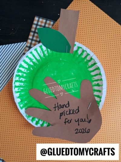 Green apple paper plate craft with a brown paper handprint cutout on top labeled Hand picked for you! 2026, perfect for an apple picking craft.