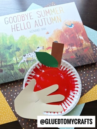 A childrens apple picking craft made with a red-painted paper plate, a brown paper stem, green paper leaf, and a beige hand cutout, displayed on a table alongside the book Goodbye Summer, Hello Autumn and colorful paper.