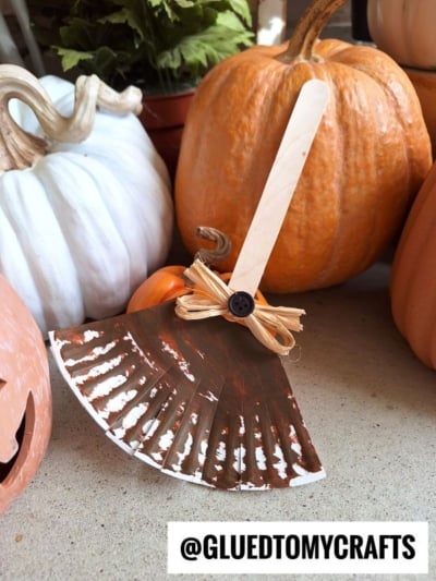 A paper plate witch broom craft made from a painted paper plate, a wooden stick, and a raffia bow with a button sits among white and orange pumpkins. Text reads @GLUEDTOMYCRAFTS.