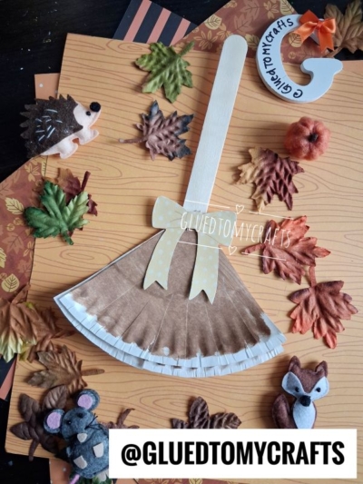 A paper plate broom craft with fake leaves, small felt animals, and autumn decor is beautifully arranged on a wood-patterned background. Text @GLUEDTOMYCRAFTS is at the bottom. This charming paper plate witch broom project perfectly captures the cozy spirit of fall.