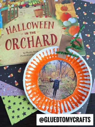 A Halloween-themed craft featuring a paper plate pumpkin picture frame with an orange-painted border displays a photo, set atop a Halloween in the Orchard book and candy corn-patterned paper. @GLUEDTOMYCRAFTS is shown at the bottom.