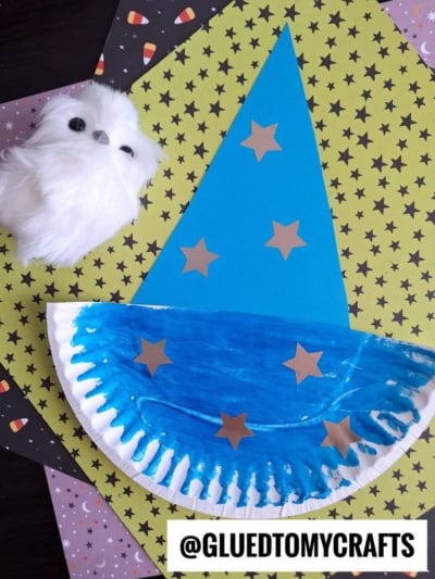 A blue paper wizard hat with gold stars sits next to a paper plate wizard hat painted blue and a white fluffy owl on star and candy corn patterned paper. Text reads @GLUEDTOMYCRAFTS.