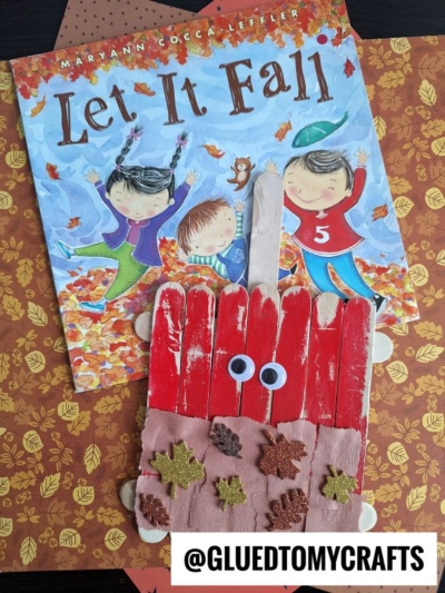 A children’s book titled Let It Fall with an autumn theme lies behind a popsicle stick craft shaped like a red rake, decorated with googly eyes, foam leaves, and a popsicle stick caramel apple on a festive fall-themed background.