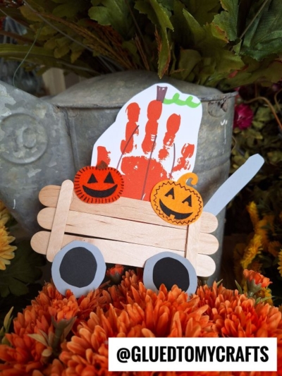 A Halloween craft featuring a popsicle stick pumpkin patch wagon with paper wheels, carrying two paper pumpkins and an orange handprint ghost, set among orange flowers. The text @GLUEDTOMYCRAFTS appears at the bottom.