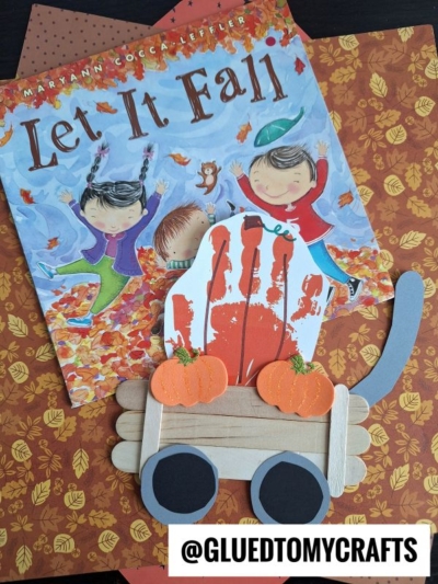 Childrens fall book Let It Fall, autumn-themed paper, and a popsicle stick pumpkin patch wagon craft featuring orange pumpkins and a red handprint, all arranged on a black surface. @GLUEDTOMYCRAFTS label at the bottom.