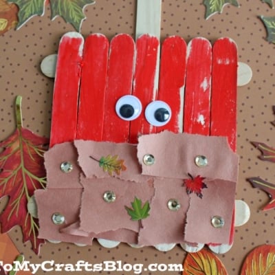 An apple craft made from red-painted popsicle sticks, like a popsicle stick caramel apple, with googly eyes, brown torn paper, sequins, stickers, and autumn leaf cutouts on a polka-dot background.