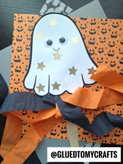 A paper ghost wand craft with star stickers, googly eyes, and a smile is on orange pumpkin-patterned paper, accented with orange and black streamers. @GLUEDTOMYCRAFTS is written at the bottom.