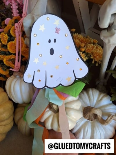A paper ghost wand decorated with gold stars and sequins is displayed among white pumpkins and yellow flowers, with colorful streamers hanging below. The Instagram handle @GLUEDTOMYCRAFTS is at the bottom.