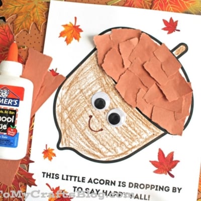 A children’s fall craft features a torn paper acorn made from brown paper pieces with googly eyes and a smile, surrounded by colorful autumn leaves.