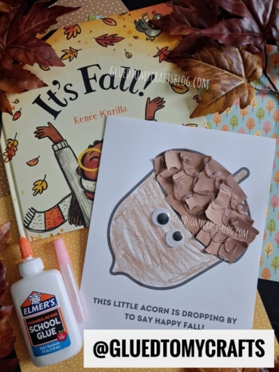A fall-themed craft setup with a children's book, autumn leaves, glue, and an adorable torn paper acorn craft made from paper and tissue. The acorn craft card says, "This little acorn is dropping by to say happy fall!.