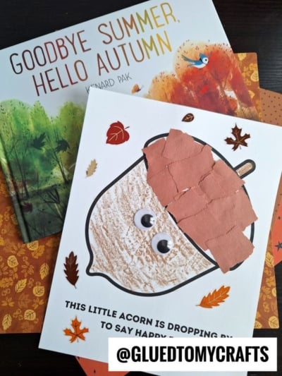 A handmade torn paper acorn card with googly eyes and a textured top sits on autumn-themed papers and a Goodbye Summer, Hello Autumn book. The card reads, This little acorn is dropping by to say happy fall! with @GLUEDTOMYCRAFTS.