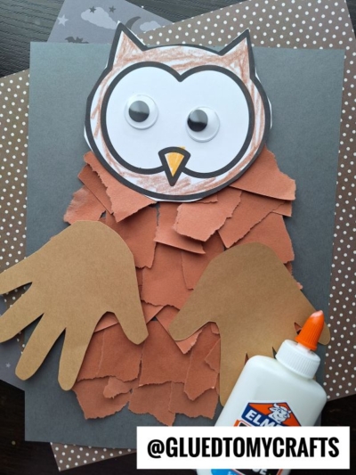 A handmade torn paper owl craft with a paper face, googly eyes, brown torn paper feathers, and handprint wings, displayed on layered patterned paper with a bottle of glue nearby. @GLUEDTOMYCRAFTS is written at the bottom.