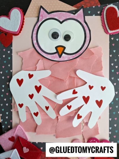 A handmade torn paper owl card features a pink owl face with googly eyes, a beak, and paper hands adorned with red hearts, set against a background of torn pink paper and surrounded by felt hearts.