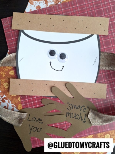 A handprint smore craft made from paper with googly eyes, a smile, and handprint cutouts reading “Love you Smore much!” Burlap ribbon acts as arms. Scrapbook papers and the @GLUEDTOMYCRAFTS label are visible.