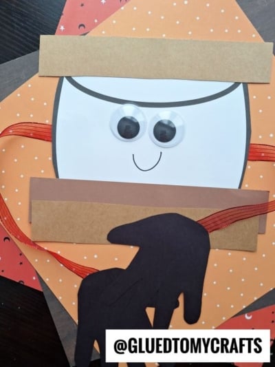 A handmade paper ninja face with googly eyes, orange and brown backgrounds, and a black paper silhouette of a goat. Red ribbon accents are included, along with a playful handprint smore detail for extra charm. @GLUEDTOMYCRAFTS text is at the bottom.