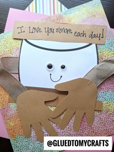 A cute handmade card features a googly-eyed, smiling handprint smore with paper arms hugging itself. Above, a sign reads I love you smore each day! @GLUEDTOMYCRAFTS.