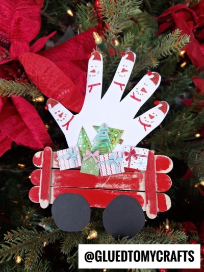 A handmade Christmas craft shaped like a popsicle stick snowman wagon, featuring a white handprint of snowmen and tiny paper presents. This festive ornament hangs on a Christmas tree adorned with red poinsettias.
