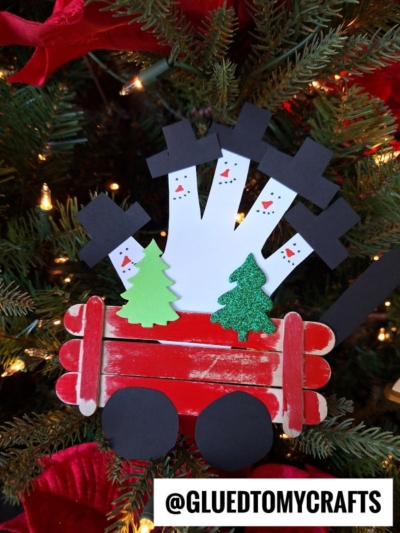 A popsicle stick snowman wagon Christmas ornament, adorned with green trees and four white handprint snowmen, hangs on a festive tree. The Instagram handle @GLUEDTOMYCRAFTS is at the bottom.