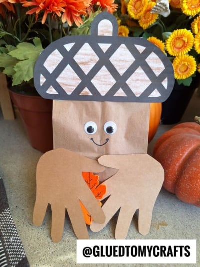 A charming paper bag acorn craft featuring googly eyes, a smiling face, and handprint cutouts holding a paper leaf. Flowers and a pumpkin decorate the background, while the @GLUEDTOMYCRAFTS text appears at the bottom.
