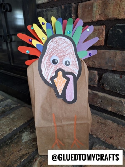 A paper bag turkey craft made from a brown paper bag, colored paper feathers, googly eyes, and a crayon-drawn face, sitting on a brick surface. @GLUEDTOMYCRAFTS is labeled at the bottom.