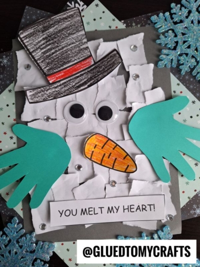 A handmade torn paper snowman craft, featuring green paper handprint arms, large googly eyes, an orange carrot nose, a black hat, and the words “YOU MELT MY HEART!” Snowflakes adorn the background.