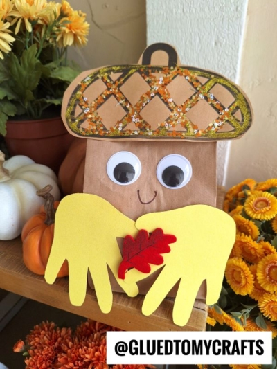 A fall-themed craft features a paper bag acorn with googly eyes, yellow handprint cutouts as arms holding a red paper leaf, surrounded by autumn flowers and pumpkins. Text reads @GLUEDTOMYCRAFTS.