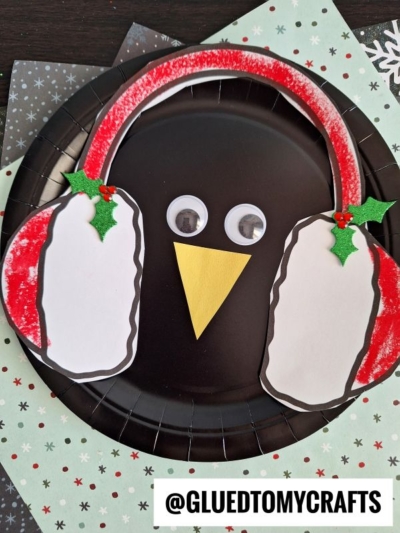 A cute ear muff penguin face made from a black paper plate with googly eyes, a yellow paper beak, and white and red earmuffs decorated with green holly, set on patterned paper. @GLUETOMYCRAFTS is written at the bottom.