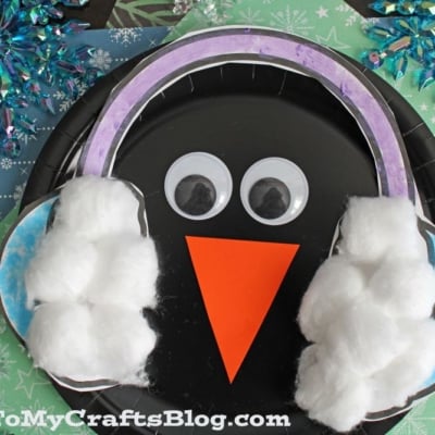 A black paper plate ear muff penguin with googly eyes, an orange paper beak, and cotton ball earmuffs sits on a blue snowflake-patterned background.