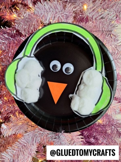 A cute ear muff penguin paper plate craft featuring googly eyes, an orange paper beak, green paper and cotton ball earmuffs, displayed against pink tinsel. The bottom includes the text @GLUEDTOMYCRAFTS.