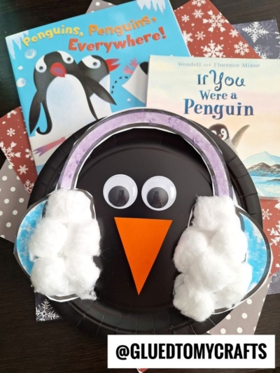 An adorable ear muff penguin face made from a black paper plate, orange beak, googly eyes, and purple earmuffs with cotton balls, surrounded by penguin-themed children’s books. Text: @GLUEDTOMYCRAFTS.