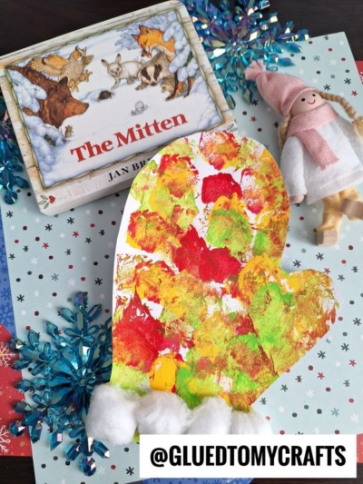 A colorful, pom pom stamped mitten with cotton ball trim is placed on winter-themed paper, next to a children’s book titled “The Mitten” and a small winter doll. Snowflake decorations are also visible.