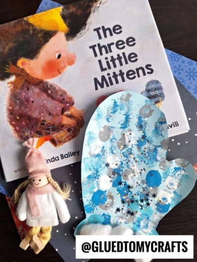 A children’s book titled “The Three Little Mittens,” a blue pom pom stamped mitten-shaped craft with snowflake stickers, and a small doll in a pink hat and scarf are arranged on a table. Text reads @GLUEDTOMYCRAFTS.
