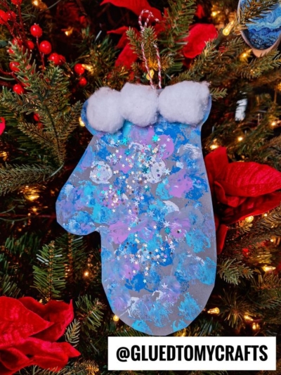 A pom pom stamped mitten-shaped ornament, decorated with blue, purple, and white paint, topped with white cotton balls and star-shaped glitter, hangs on a Christmas tree with red flowers. Text at the bottom reads @GLUEDTOMYCRAFTS.