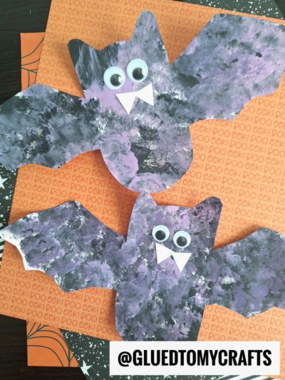 Two handmade pom pom stamped bats with purple, black, and white painted wings, large googly eyes, and white paper fangs are displayed on an orange patterned background. @GLUEDTOMYCRAFTS is printed at the bottom.
