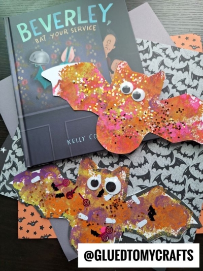 Two colorful, glittery pom pom stamped bat crafts with googly eyes are displayed on bat-patterned paper alongside the book Beverley, Bat Your Service by Kelly Cook. Text at the bottom reads @GLUEDTOMYCRAFTS.