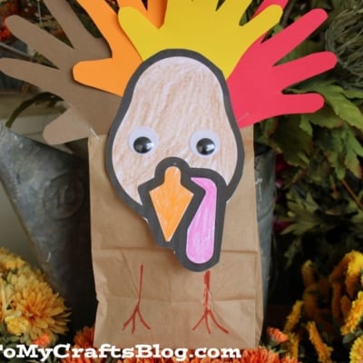 A paper bag turkey craft featuring colorful handprint feathers, googly eyes, and a drawn beak and wattle, surrounded by autumn flowers.