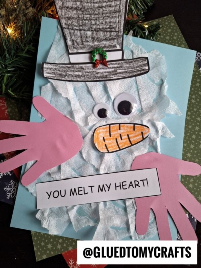 A handmade torn paper snowman craft with tissue paper for the body, large pink paper hands, and a sign that says You melt my heart! on blue paper. The hat features a tiny wreath and there’s an @gluedtomycrafts label at the bottom.