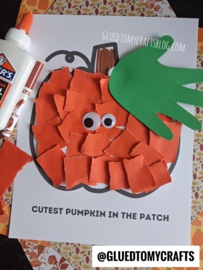 A kids’ torn paper pumpkin craft made of vibrant orange paper on a pumpkin outline with googly eyes, a green handprint as a leaf, Elmer’s glue, and the text “CUTEST PUMPKIN IN THE PATCH.” Website watermark is also visible.