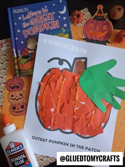 A torn paper pumpkin children’s craft made with vibrant orange scraps, glitter, and a green handprint is on display, accompanied by the book Letters to the Great Pumpkin, glue, and playful pumpkin-themed decorations.