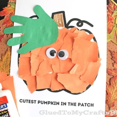 A torn paper pumpkin craft made from ripped orange paper and a green handprint stem, glued to a pumpkin outline with googly eyes and the text CUTEST PUMPKIN IN THE PATCH. A glue bottle sits beside this adorable creation.