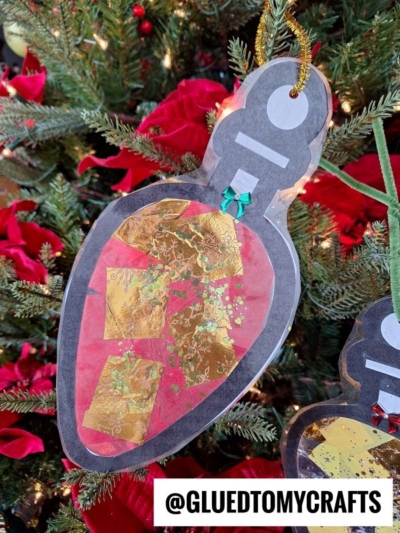 A homemade Christmas light suncatcher ornament, shaped like a bulb and decorated with red and gold tissue paper, hangs on a tree adorned with red flowers and lights. The handle @GLUEDTOMYCRAFTS appears at the bottom of the image.
